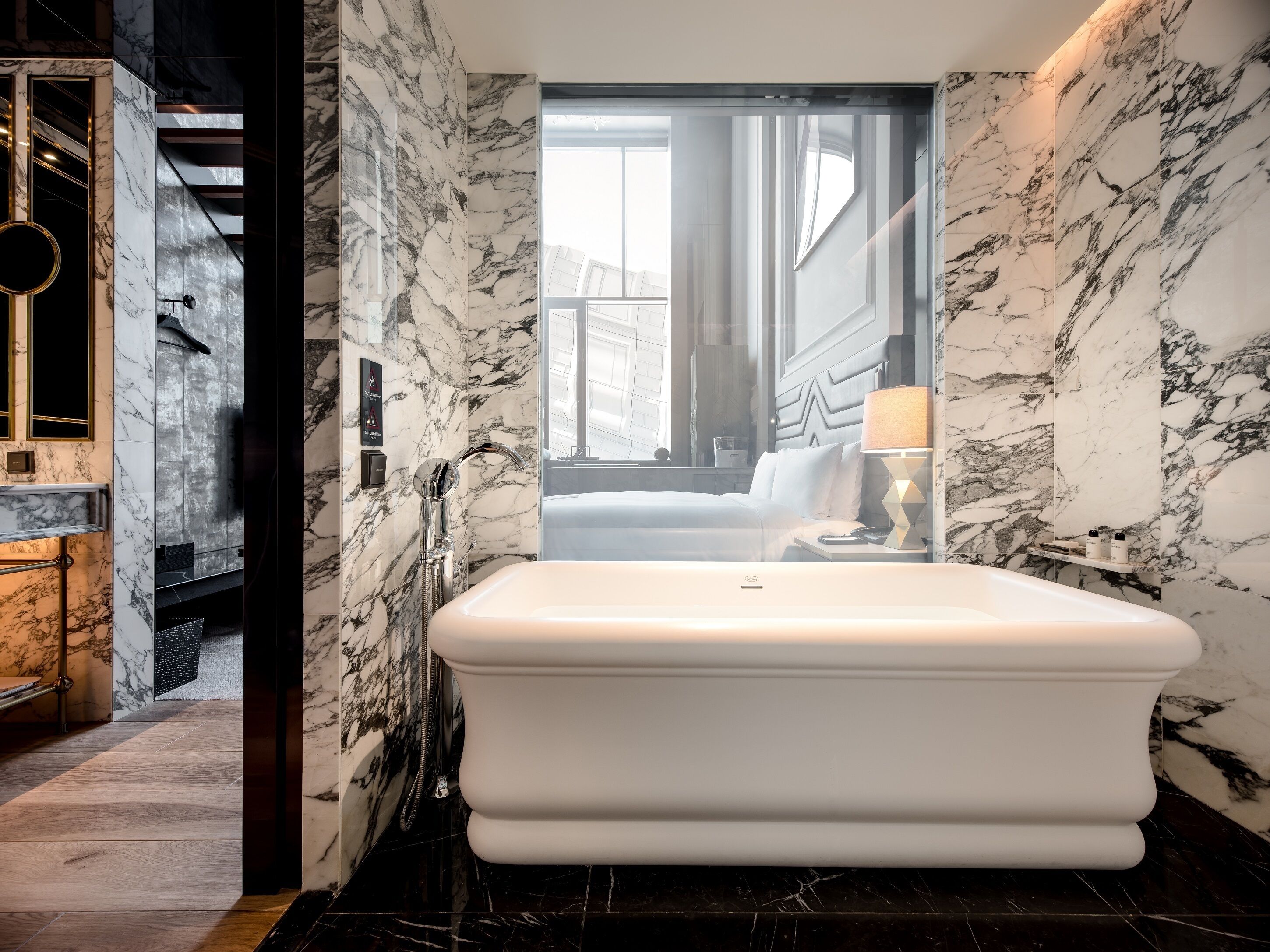A modern bathroom with a large white bathtub, marble walls, and a view of a cityscape through a large window.