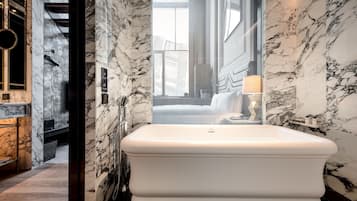 A modern bathroom with a large white bathtub, marble walls, and a view of a cityscape through a large window.
