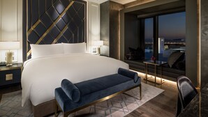 Een moderne hotelkamer met een groot bed, een bankje, een nachtkastje, een lamp, uitzicht op de stad in de schemering en een raam met een stadsgezicht.