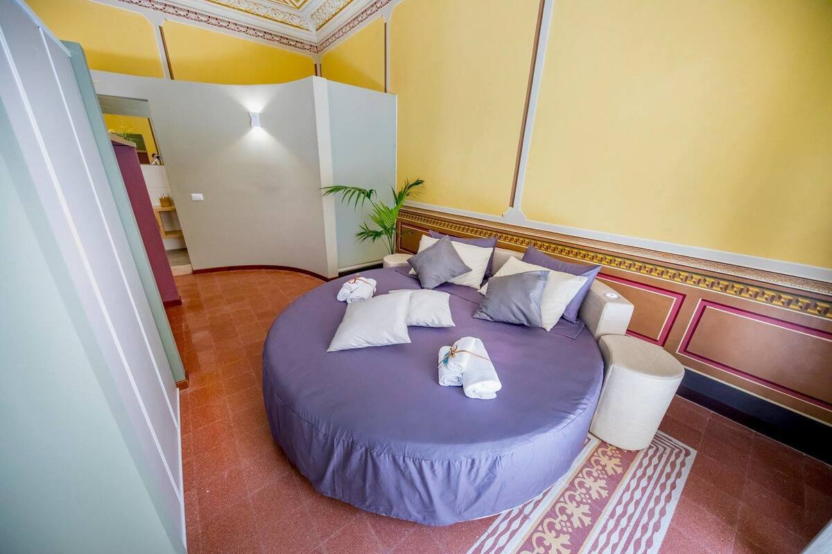 Een rond bed met een paars dekbed, meerdere kussens en een witte nachtkastje met een lamp.