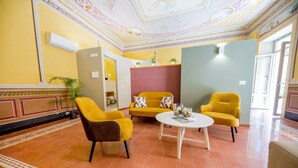 A hotel lobby with yellow walls, a patterned floor, and a ceiling with intricate designs. There are two yellow armchairs and a round table with decorative items. - Pepito Palace (Cefalù)