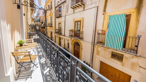 A narrow street with buildings on both sides, featuring balconies with plants and a striped awning. - Pepito Palace (Cefalù)