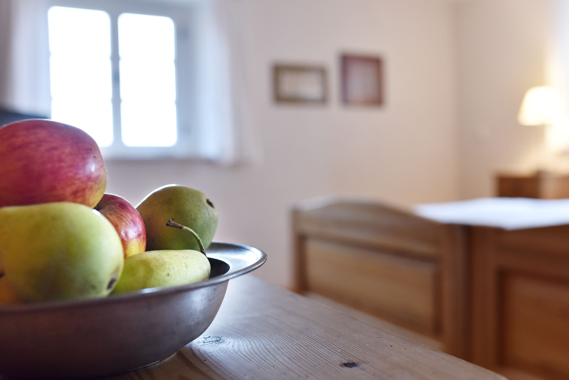 Un frutero con fruta sobre una mesa de madera en una habitación con ventana y una cama al fondo.