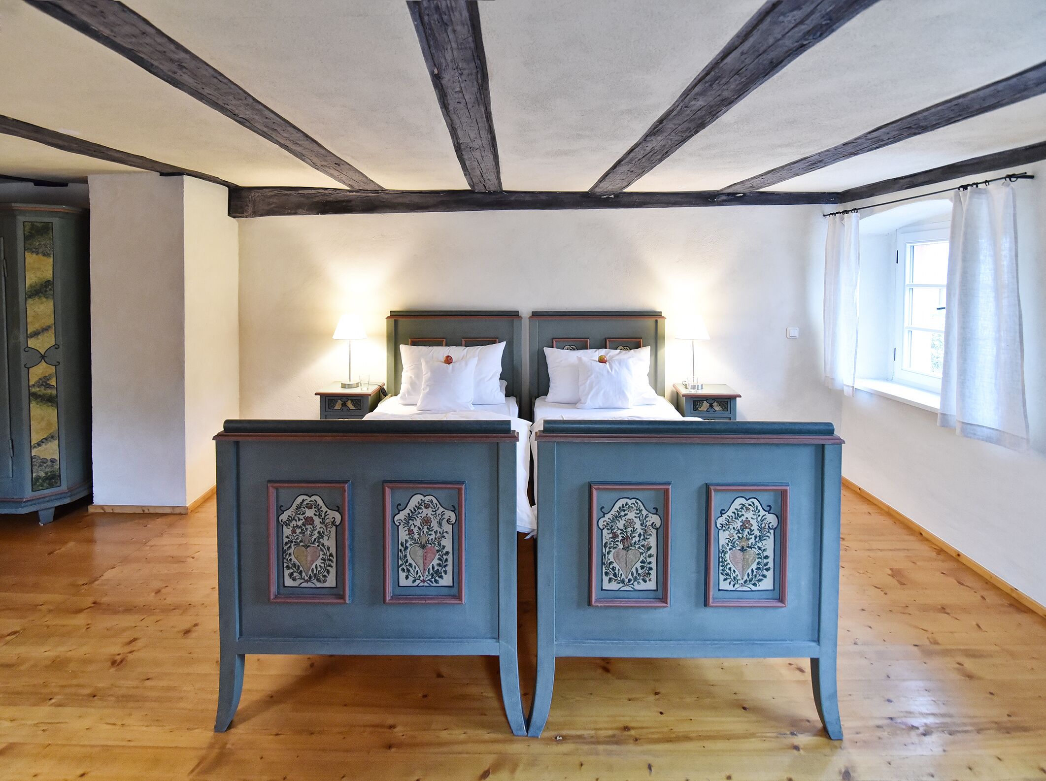 A room with two beds, wooden floors, and a window with curtains.