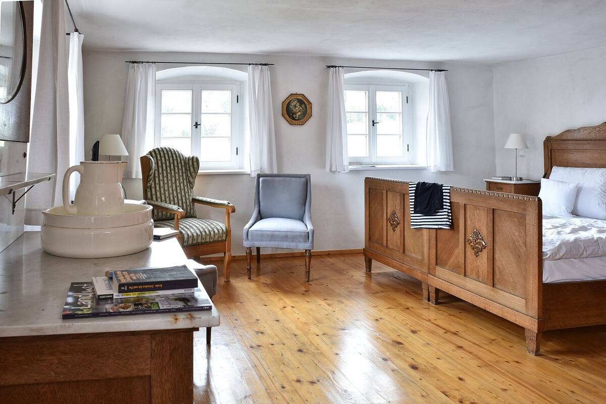 A bedroom with a wooden bed, a nightstand, a chair, a desk with a lamp, and two windows with curtains.
