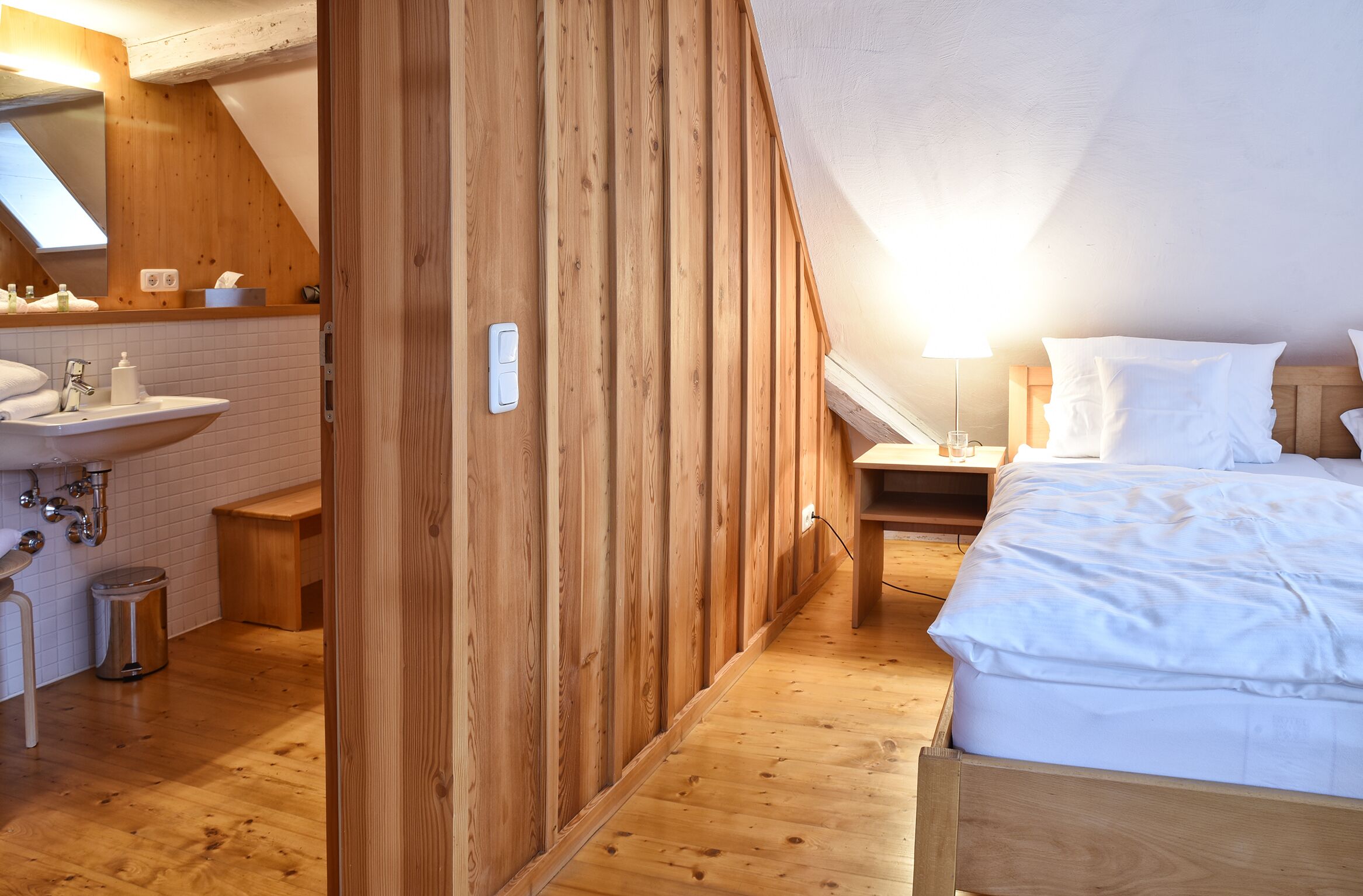 Un dormitorio con cabecera de madera, una mesita de noche, una lámpara y una colcha blanca.
