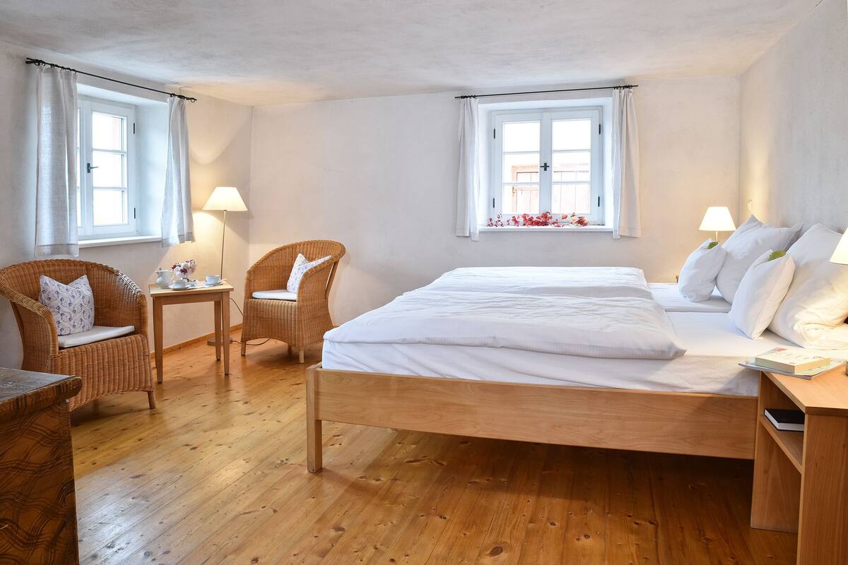 A bedroom with a wooden bed, two wicker chairs, a small table, and two windows with curtains.
