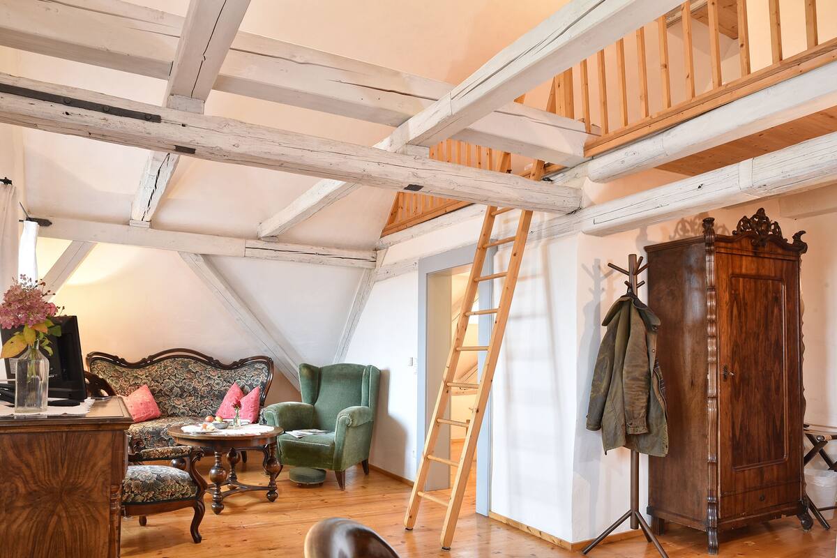 A loft bedroom with exposed wooden beams, a green armchair, a wooden cabinet, and a ladder leading to a lofted area.