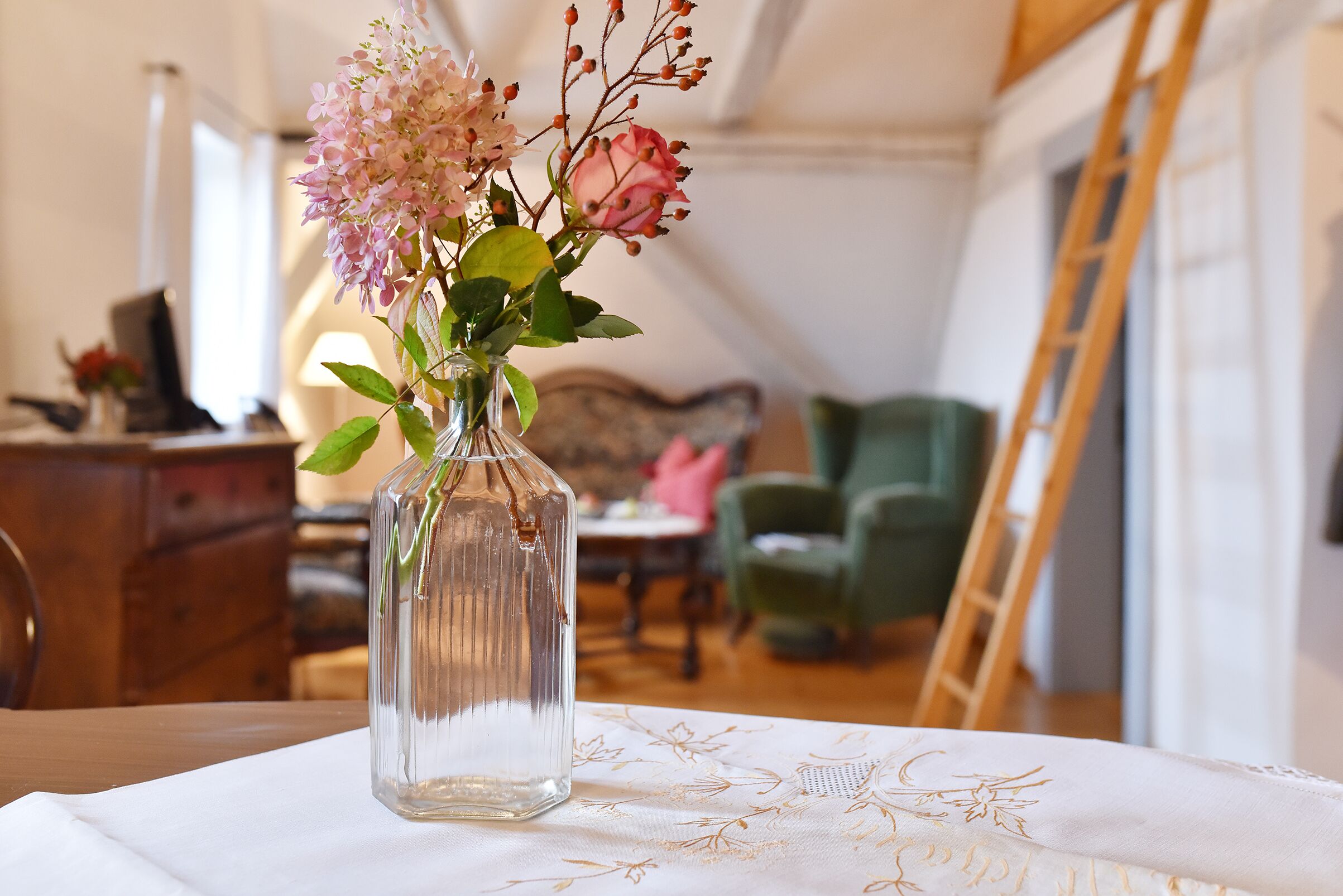Un florero de vidrio transparente con flores rosadas sobre una mesa en una habitación con piso de madera, un sillón verde y una escalera pegada a la pared.