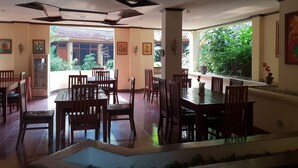 A dining area with wooden tables and chairs, a refrigerator, and a view of a garden through large windows. - Hotel Dequr Bandung (Bandung)