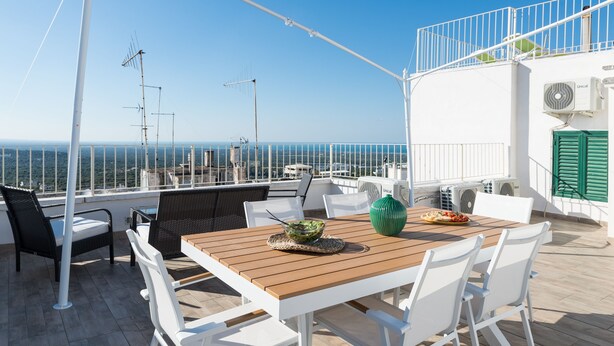 A terrace with a wooden table, white chairs, and a view of the sea. - Isabella Apartments by Wonderful Italy (Ostuni)