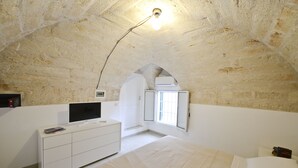 A bedroom with a stone ceiling, a bed, a TV on a white dresser, and a window with a view. - Isabella Apartments by Wonderful Italy (Ostuni)