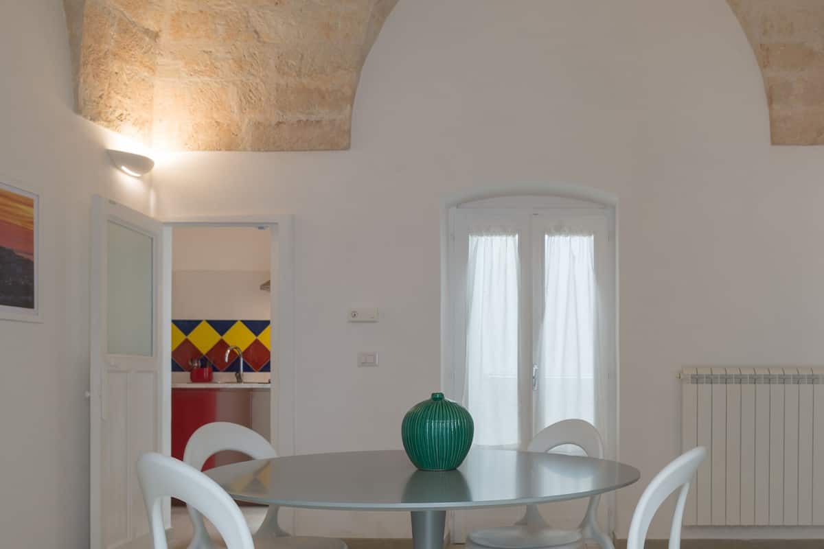 A dining area with a round table, white chairs, a green vase, and a white door with a window.