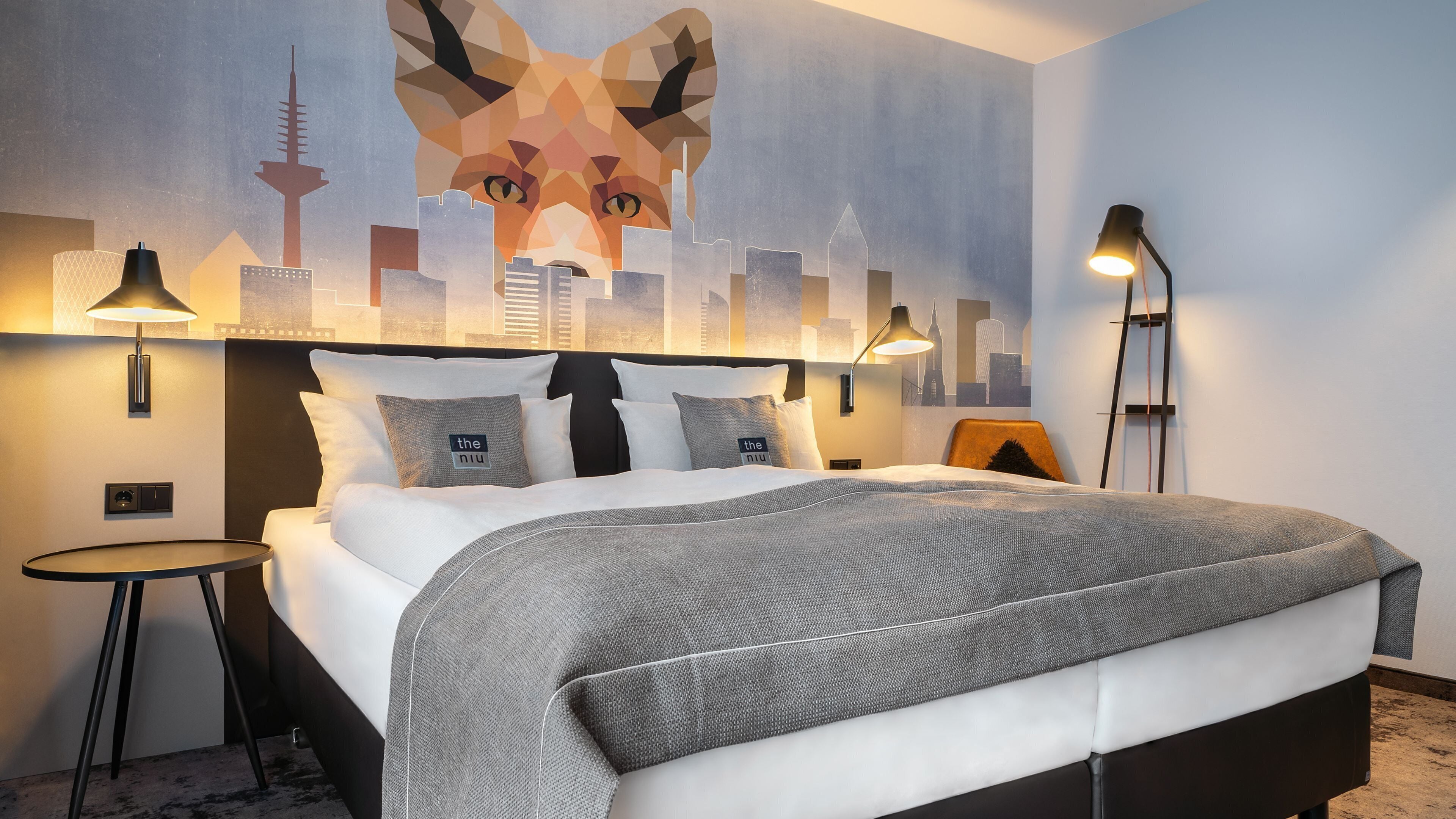 A modern hotel room with a large bed, two bedside tables, a cityscape mural, and a city skyline view.