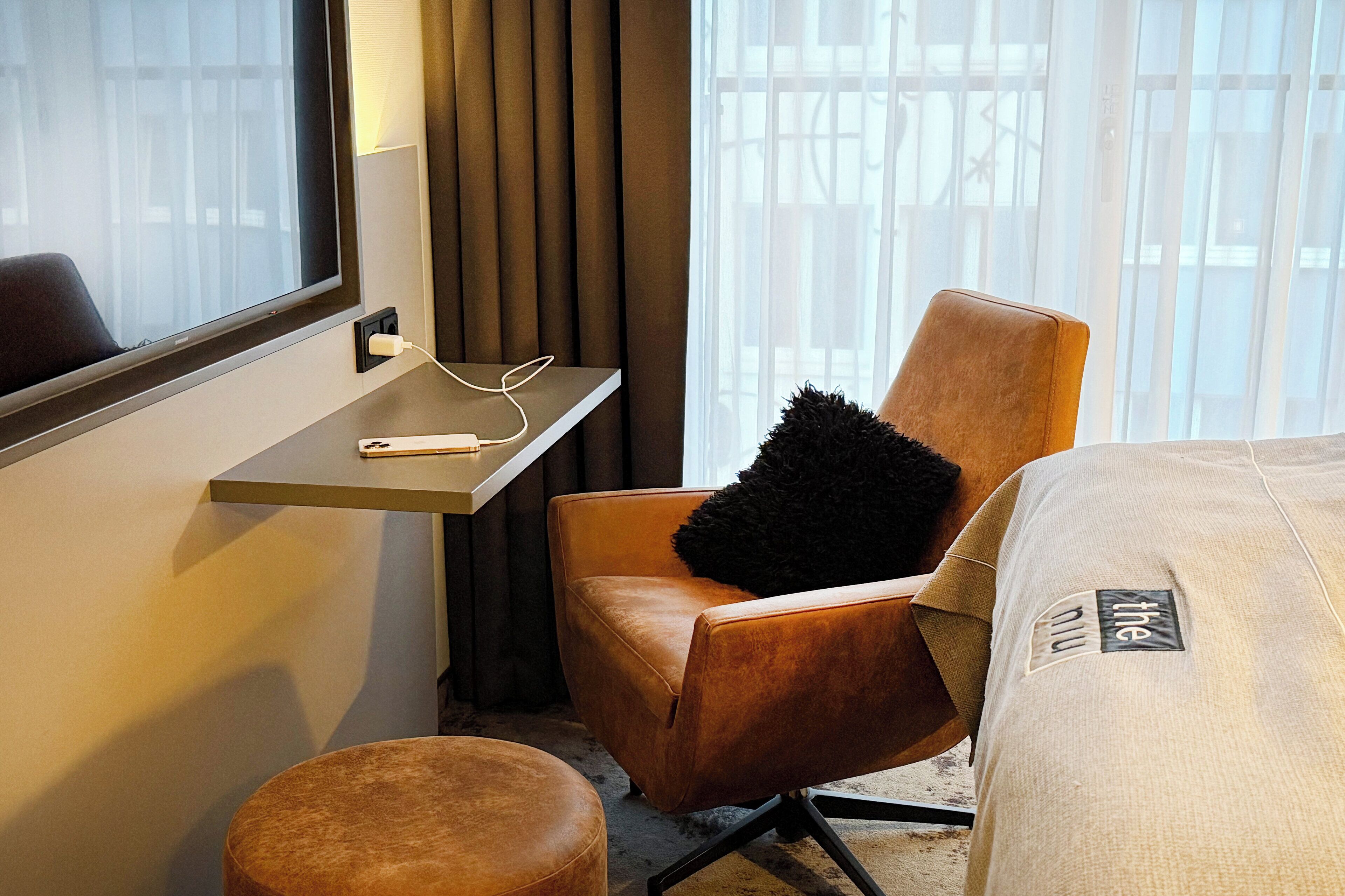 A hotel room with a single brown leather chair, a small bedside table with a white charger, and a large window with curtains.
