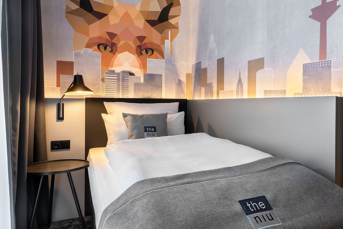 Hotellihuone, jossa on sänky, yöpöytä, lamppu ja seinällä kaupunkimaisemaa esittävä seinämaalaus.