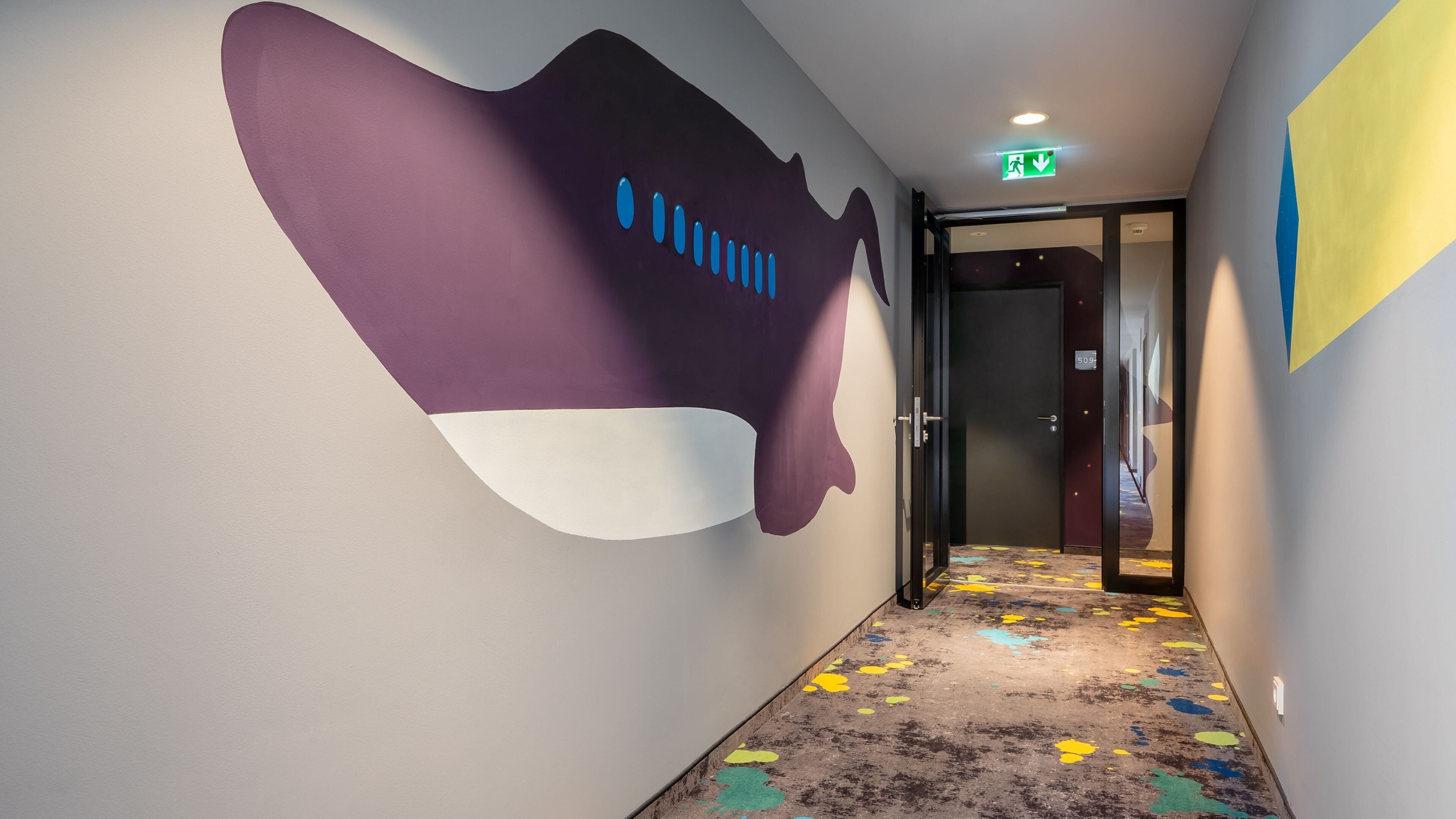 A hallway with a purple and white wall mural, a colorful carpet, and a door with a number sign.