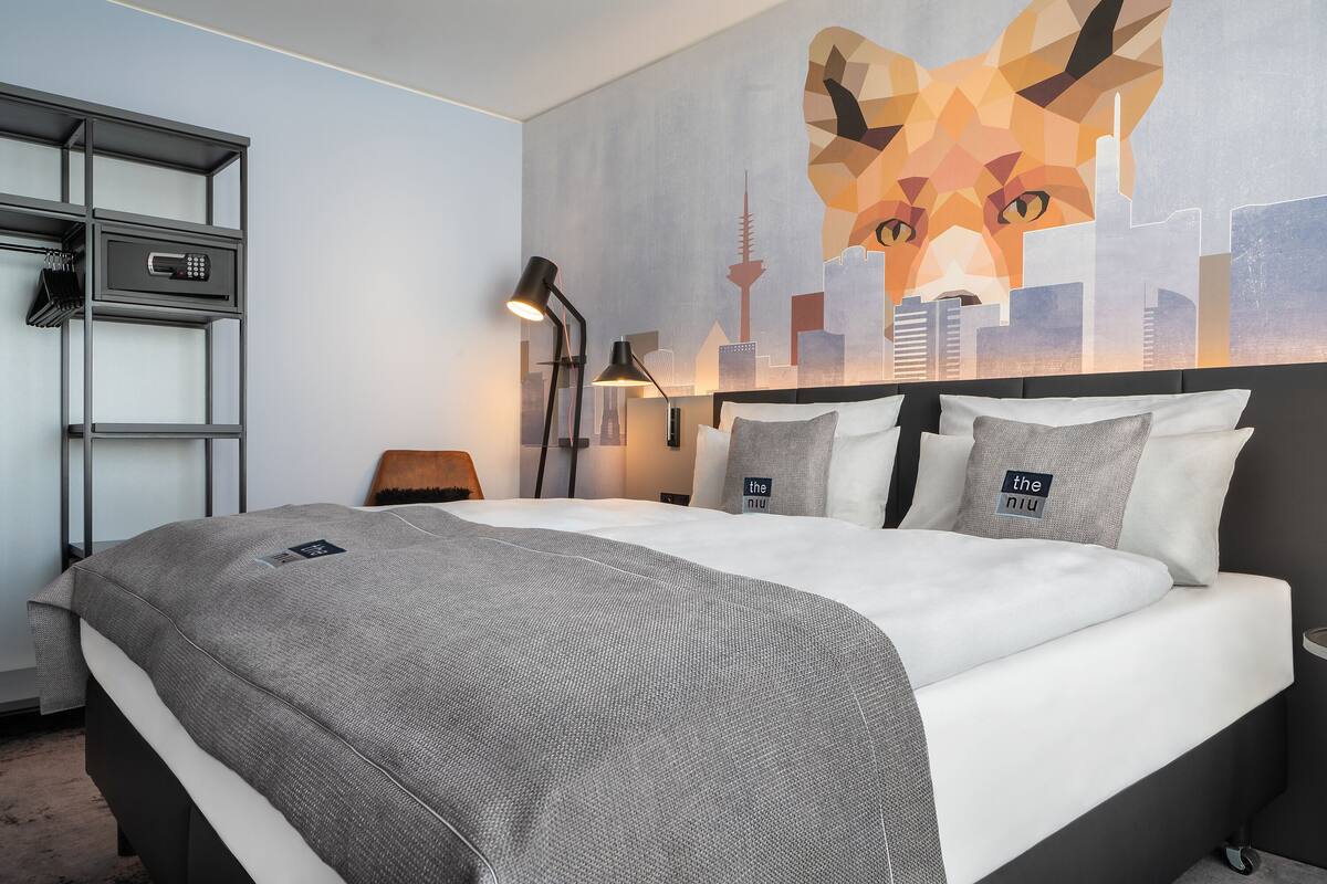 Hotellihuone, jossa on sänky, kaupunkimaisemaa esittävä seinämaalaus, työpöytä lampun kanssa ja latausasema.