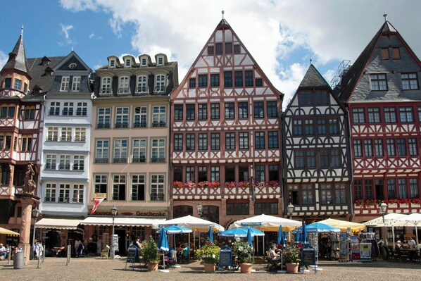 A row of traditional half-timbered buildings with a cobblestone square in front, featuring outdoor seating and flower arrangements.