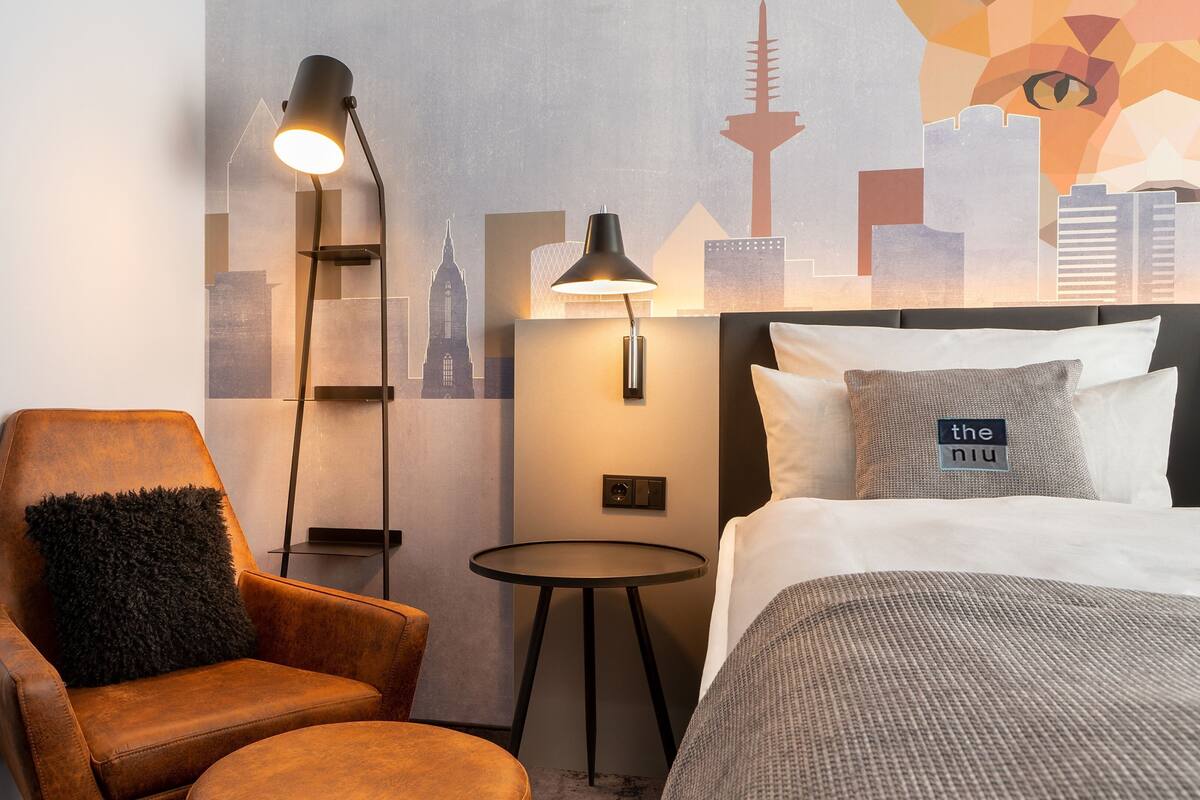 Hotellihuone, jossa on sänky, tuoli, pieni pöytä ja seinällä kaupungin siluettia esittävä taideteos.