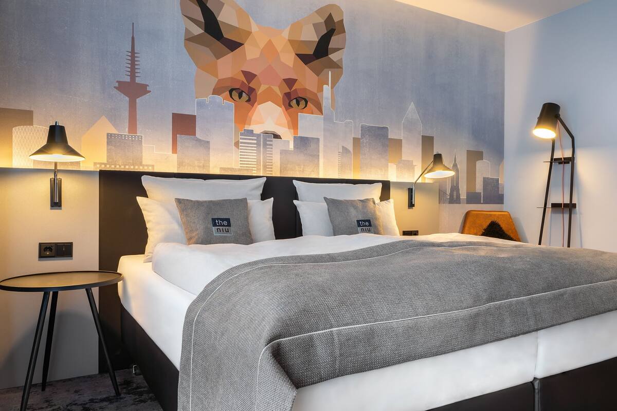 Moderni hotellihuone, jossa on suuri sänky, kaksi yöpöytää, kaupunkimaisemaa esittävä seinämaalaus ja näkymä kaupungin siluettiin.