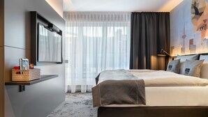 Una habitación de hotel moderna con una cama grande, un televisor montado, un mural con vista a la ciudad y vistas urbanas a través de cortinas translúcidas.