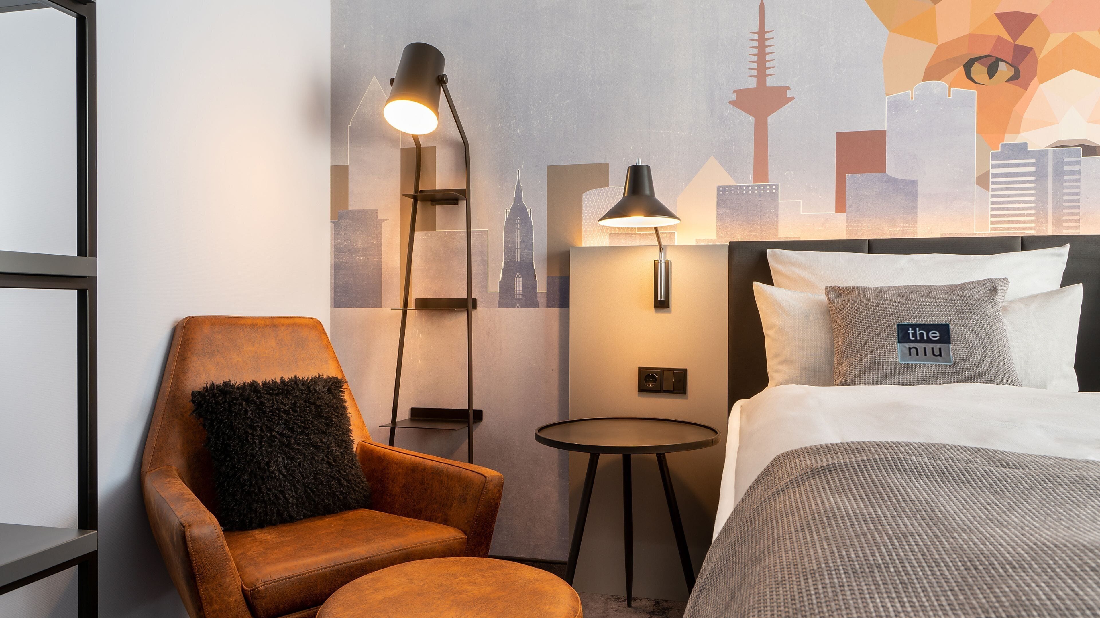 Habitación de hotel con una cama, una silla, una mesita y un cuadro de la ciudad.