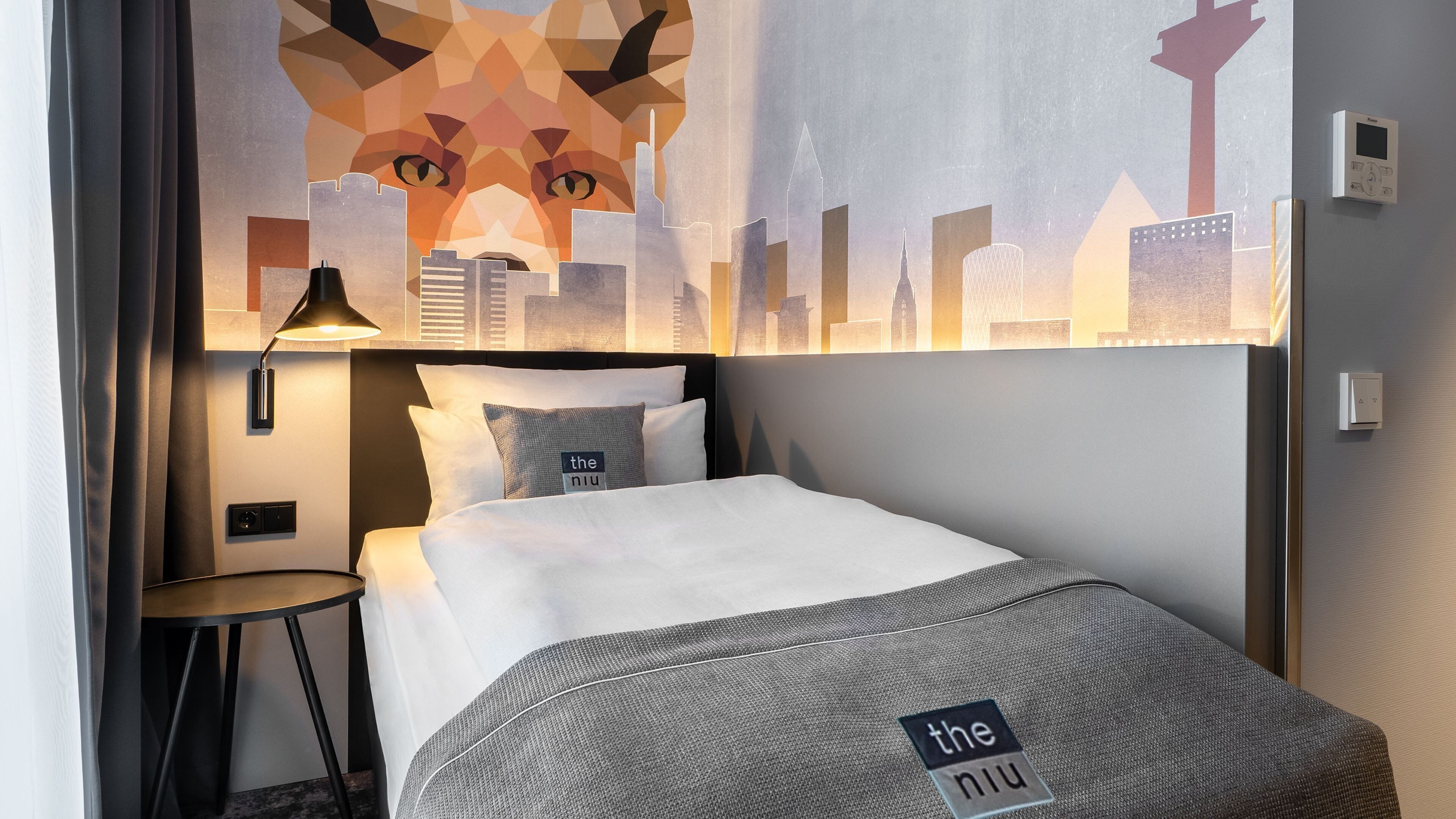 A hotel room with a bed, a nightstand, a lamp, and a cityscape mural on the wall.