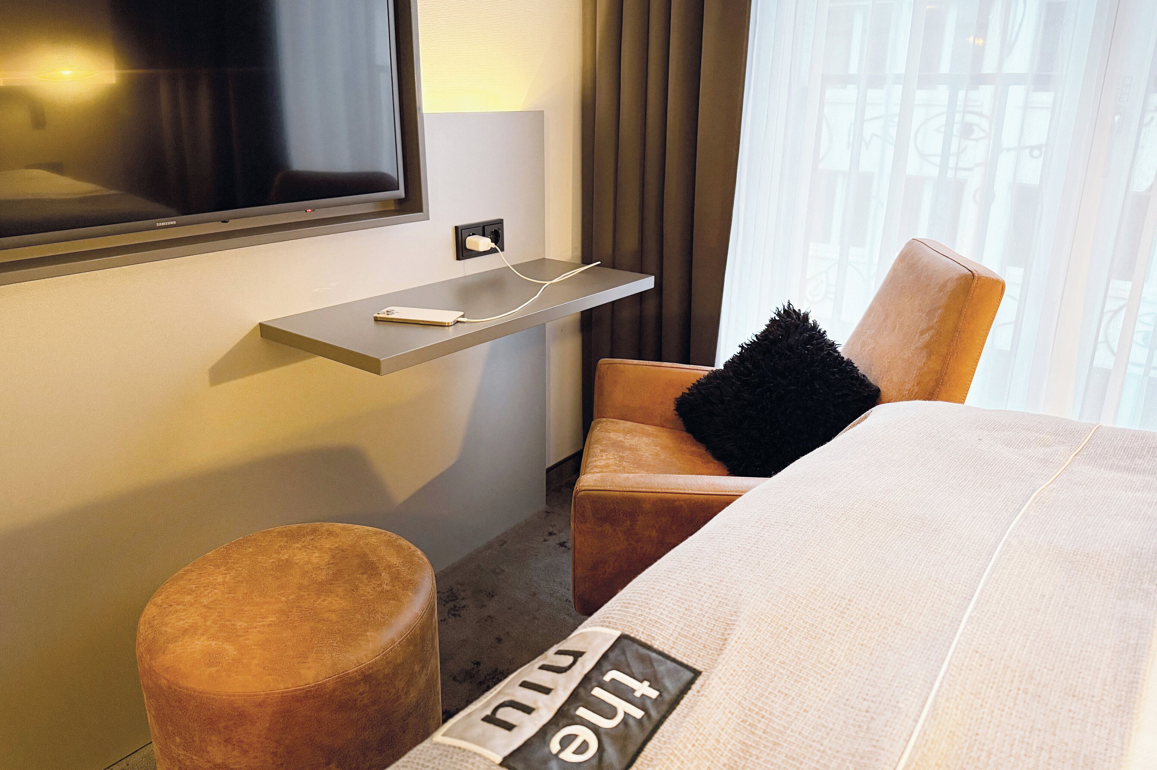 Une chambre d’hôtel avec un lit, un téléviseur fixé au mur, une petite table avec une prise électrique, un pouf marron et une chaise avec un coussin noir.