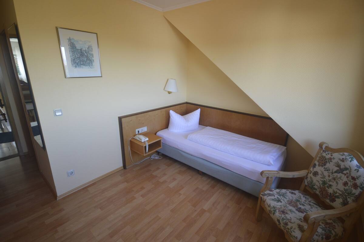 A small, single-bed room with a wooden floor, a nightstand with a lamp, and a chair.