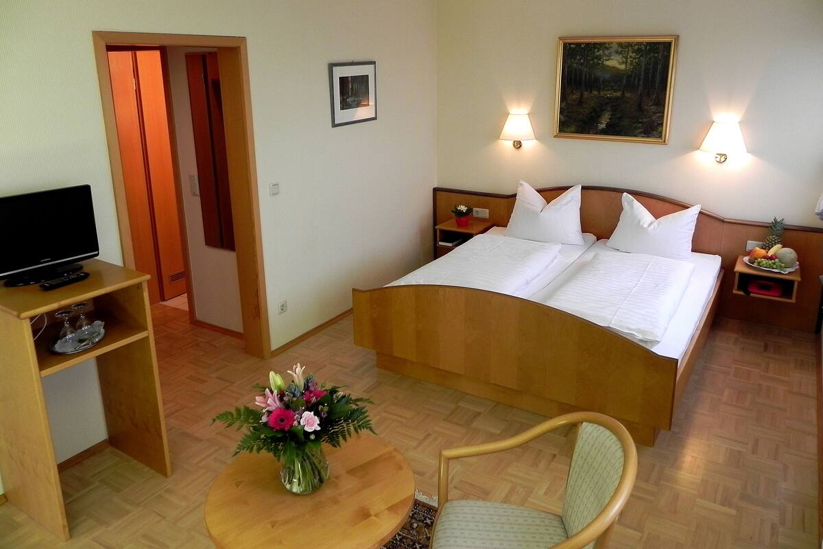 A hotel room with a bed, a TV on a stand, a chair, a small table with a vase of flowers, and a painting on the wall.