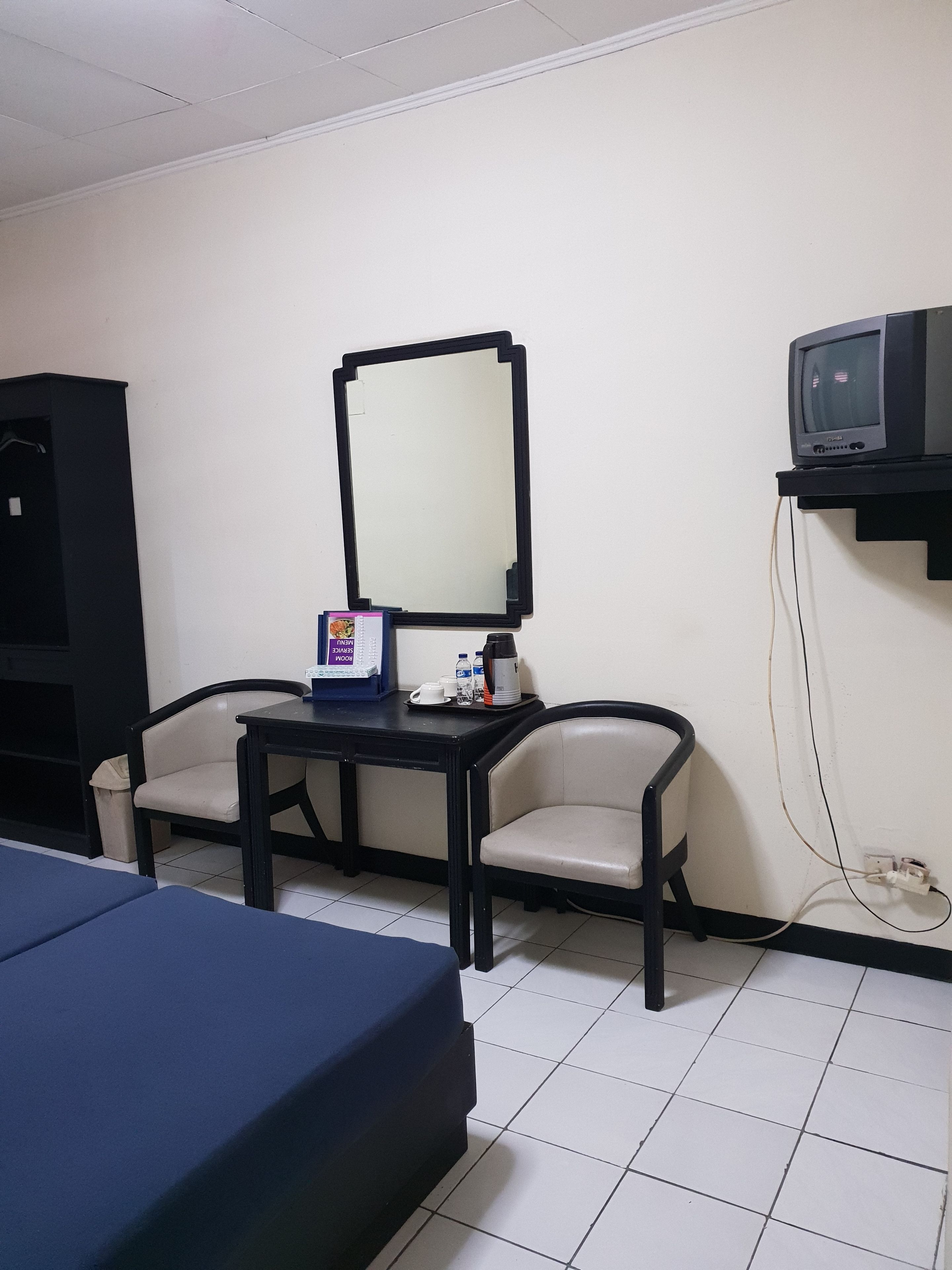 Una habitación pequeña con una cama, un escritorio con espejo, dos sillas, un televisor sobre un mueble bajo y un cubo de basura.
