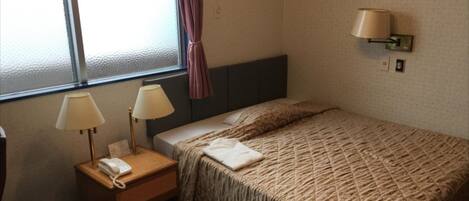 A hotel room with a bed, two bedside lamps, a telephone, a window with a curtain, and an air conditioning unit.