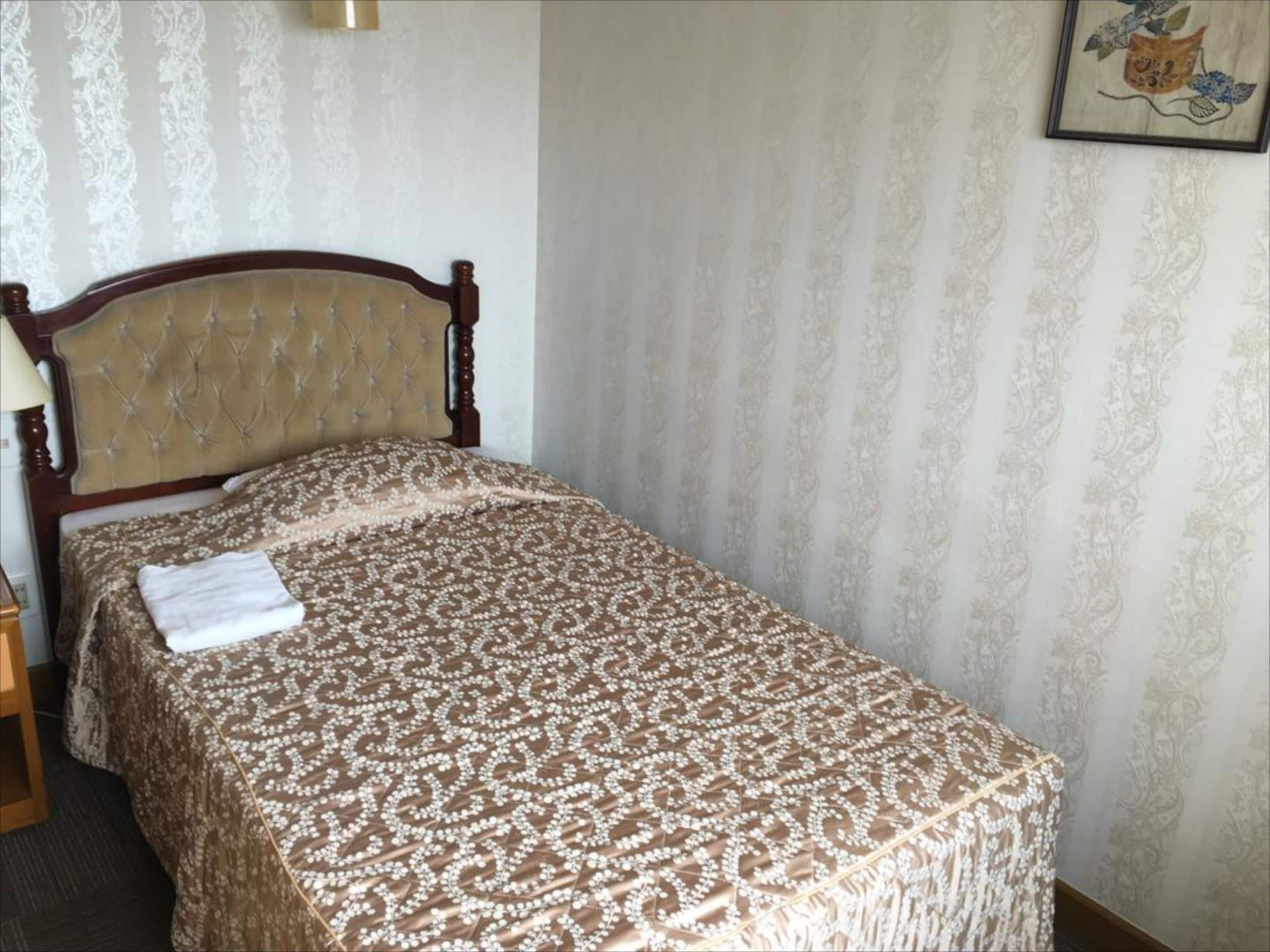 Un lit bien fait, avec une couvre-lit à motifs et une serviette pliée, une table de chevet en bois avec une lampe, et un tableau encadré au mur.