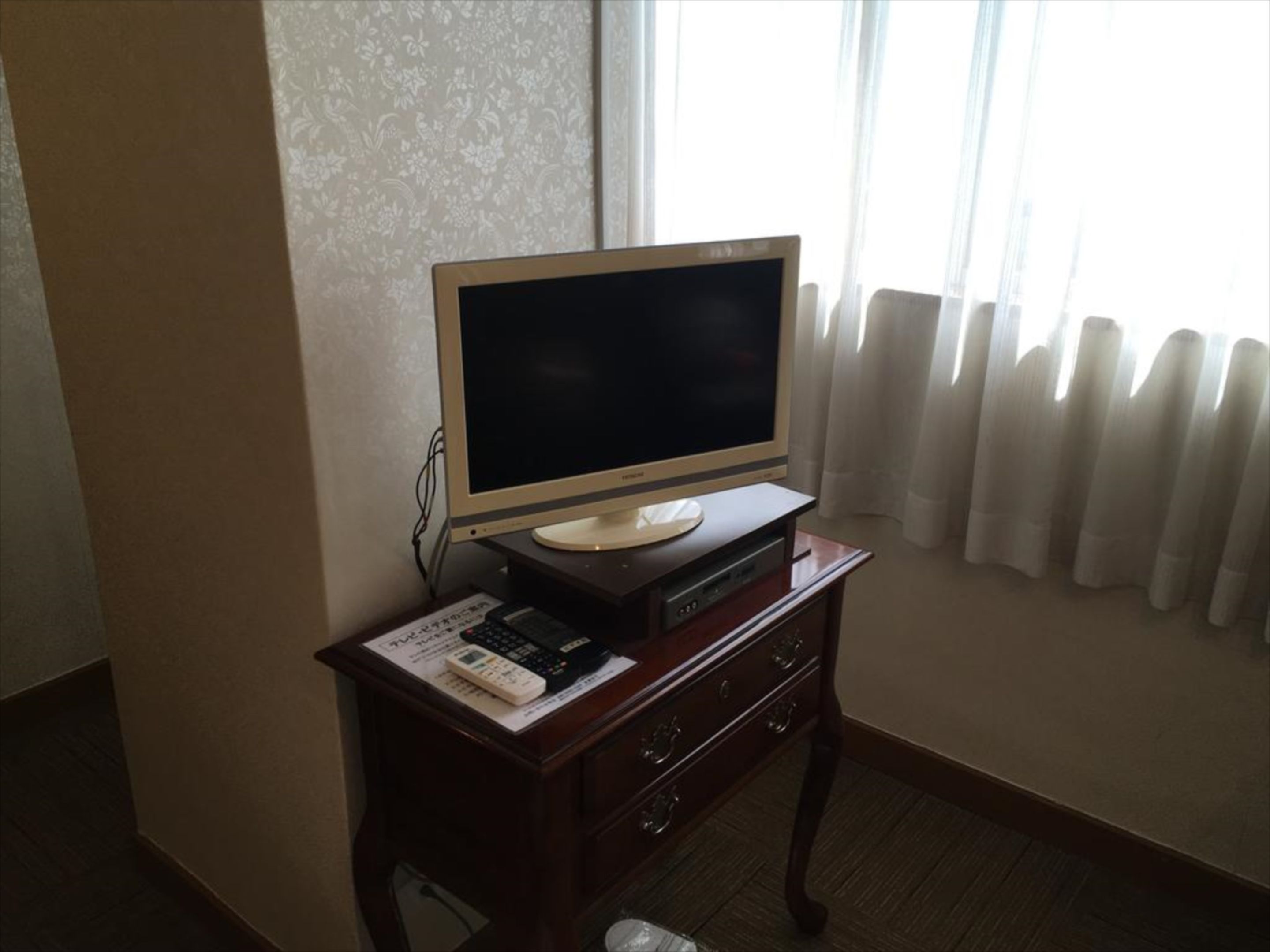 A television on a wooden cabinet in a room with a patterned wallpaper and a window with curtains.