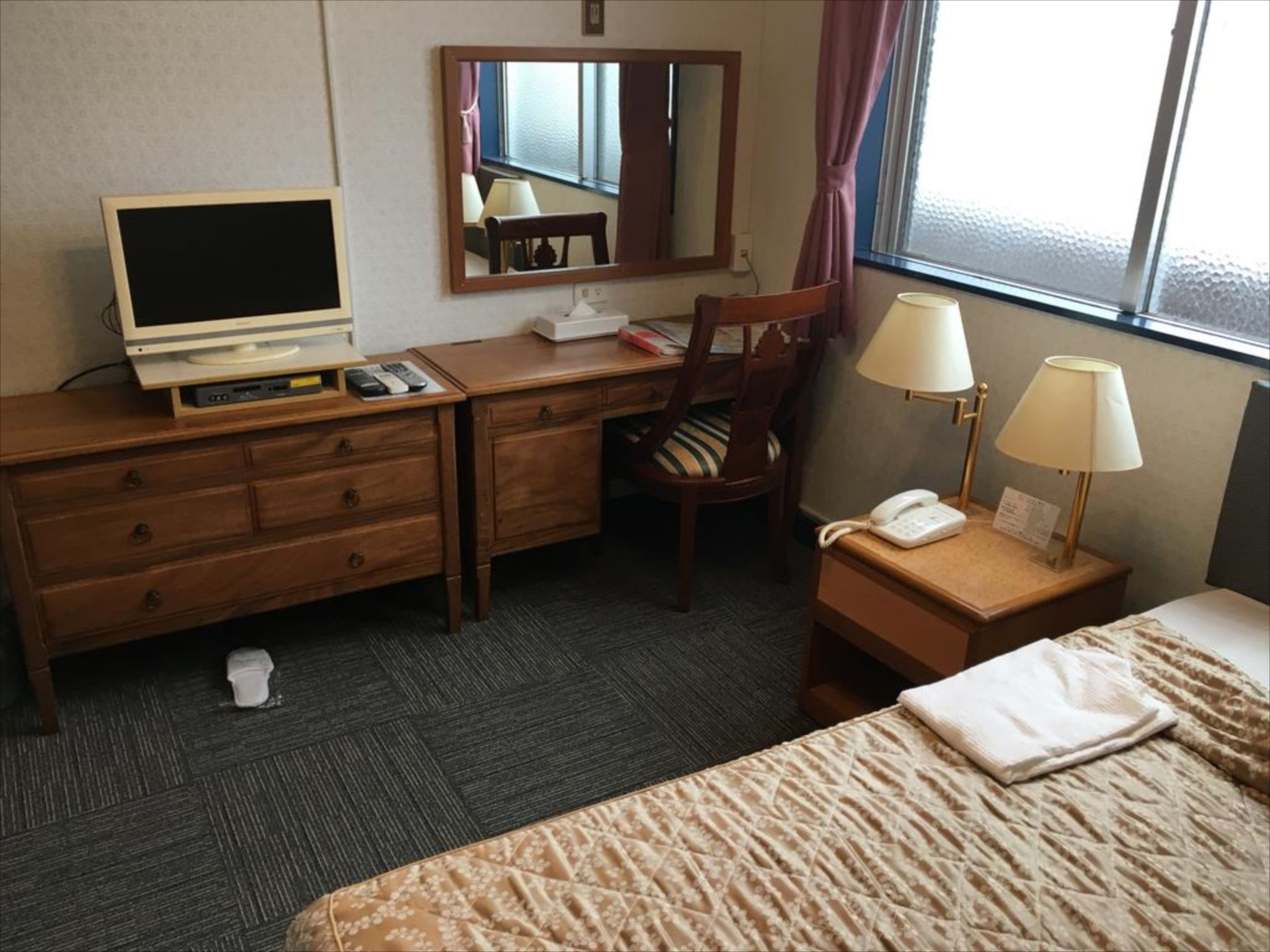 A hotel room with a bed, a desk with a laptop, a chair, a bedside table with a lamp, and a window with curtains.