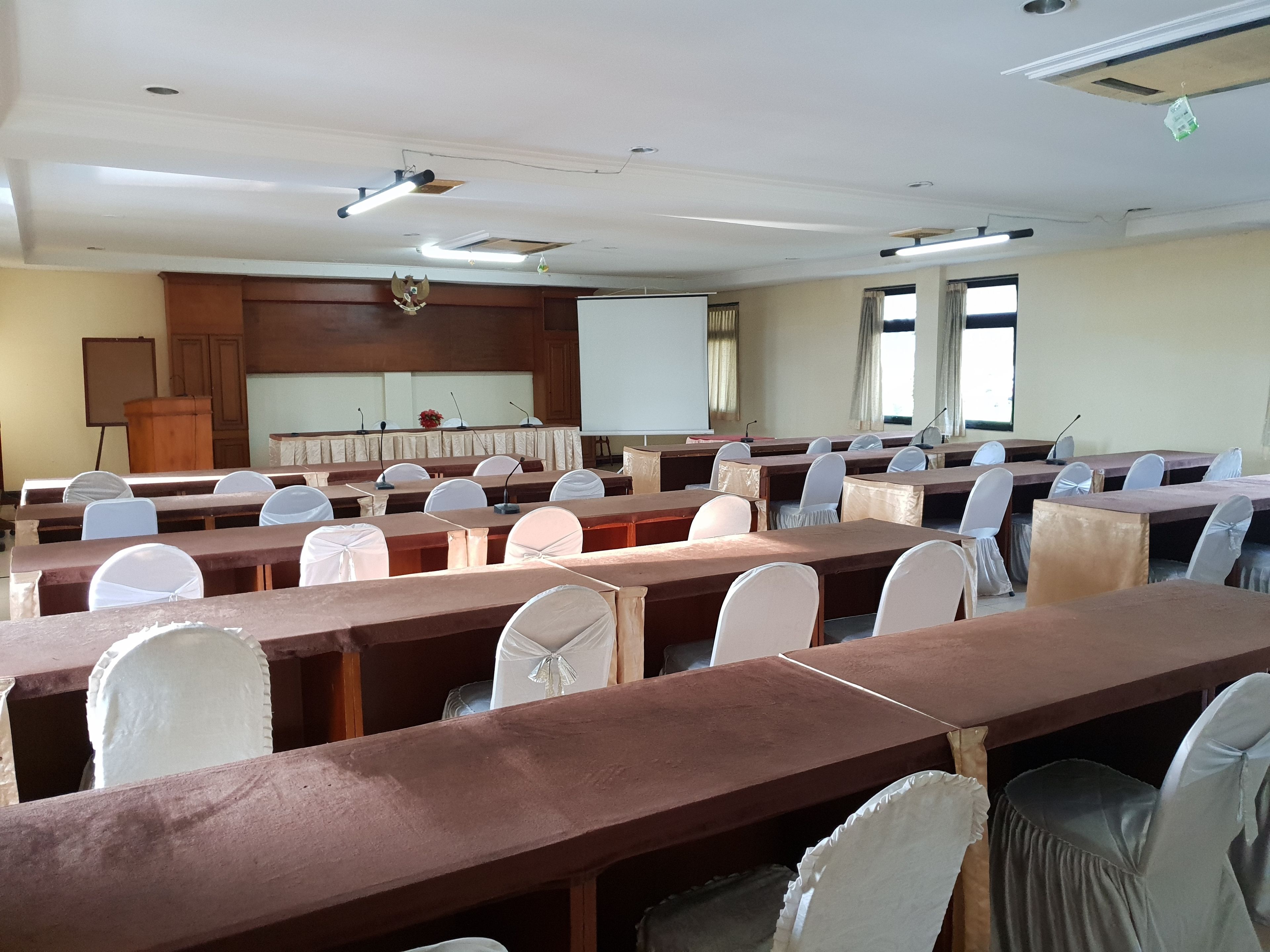 Un salón de conferencias con largas mesas, sillas, un atril, una pantalla de proyector y una pizarra.