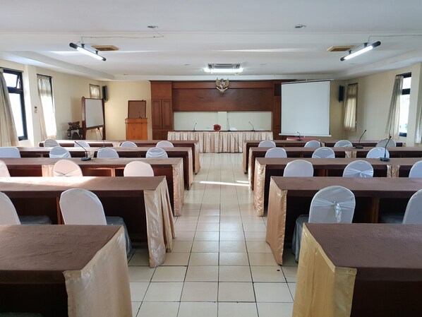 Un salón de conferencias con filas de mesas y sillas, un atril, una pizarra y una pantalla de proyector.