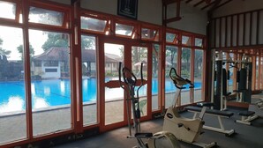 Un gimnasio con amplios ventanales que dan a una piscina, equipamiento de ejercicio y vistas a un edificio con tejado de tejas.