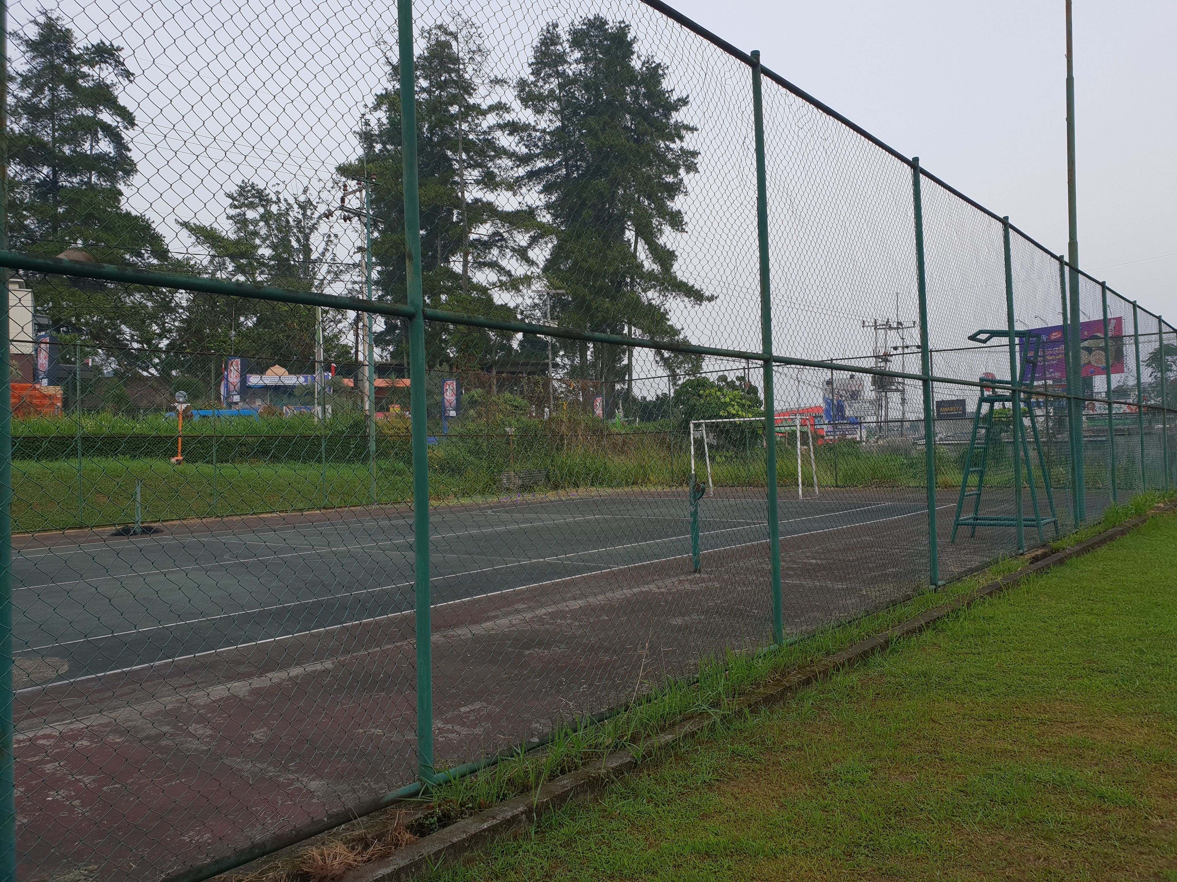 Una cancha deportiva cercada con estructura metálica verde, superficie de concreto y zona de césped adyacente.