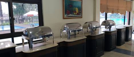 A buffet setup with multiple self-service stations, a painting on the wall, and a view of a swimming pool outside.
