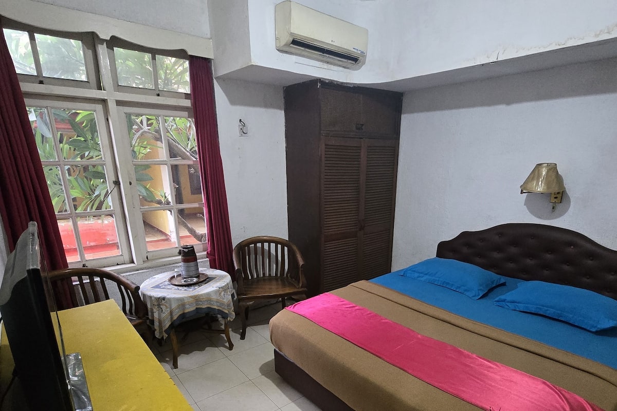 A bedroom with a bed, a table, a chair, an air conditioner, and a window with curtains.
