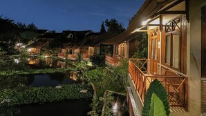 Une rangée de chalets en bois dotés de balcons donnant sur un étang paisible, le tout entouré d’une végétation luxuriante.