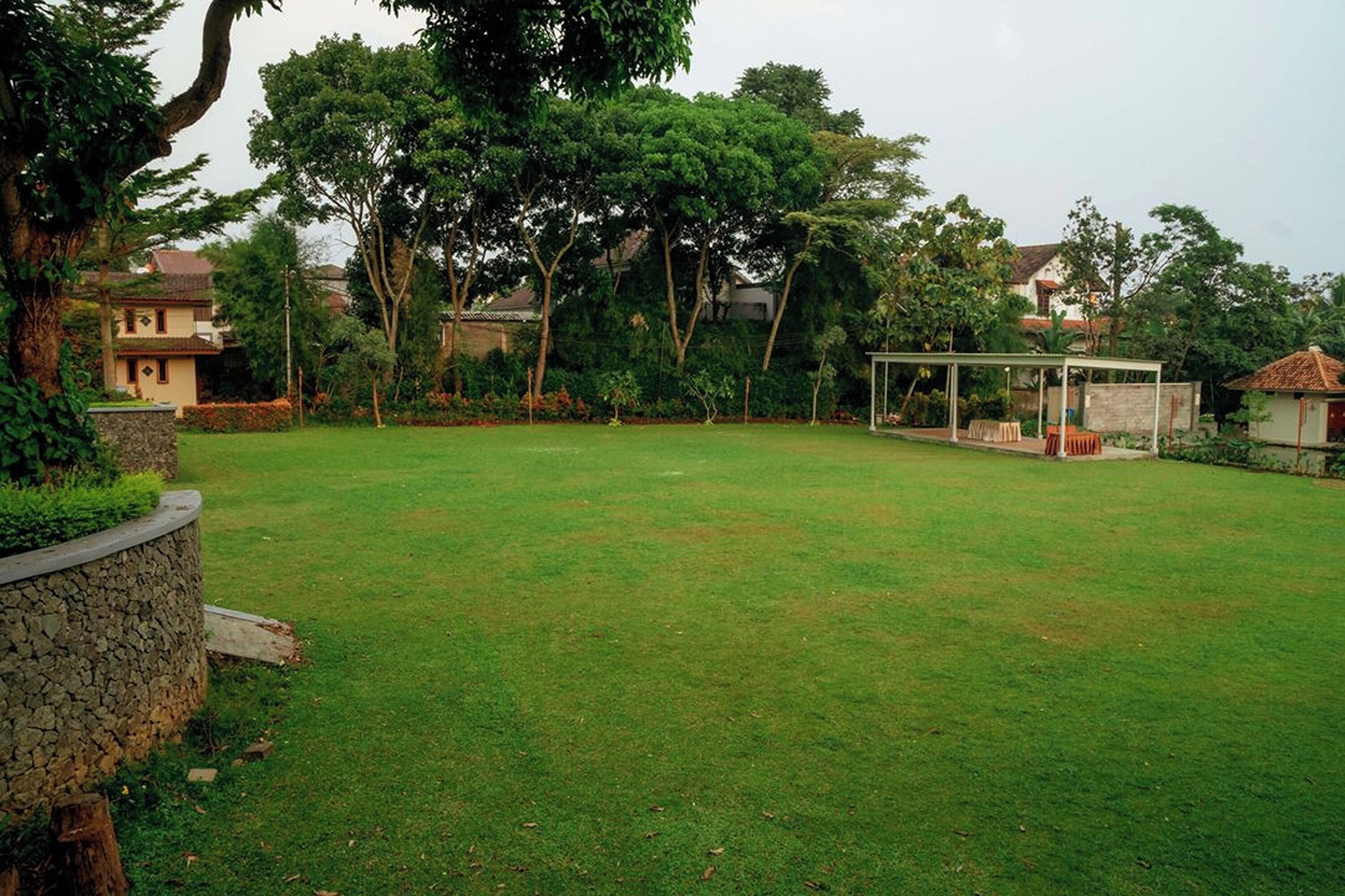 Un patio trasero cubierto de césped con un muro de piedra de contención, un área de estar techada y árboles circundantes.