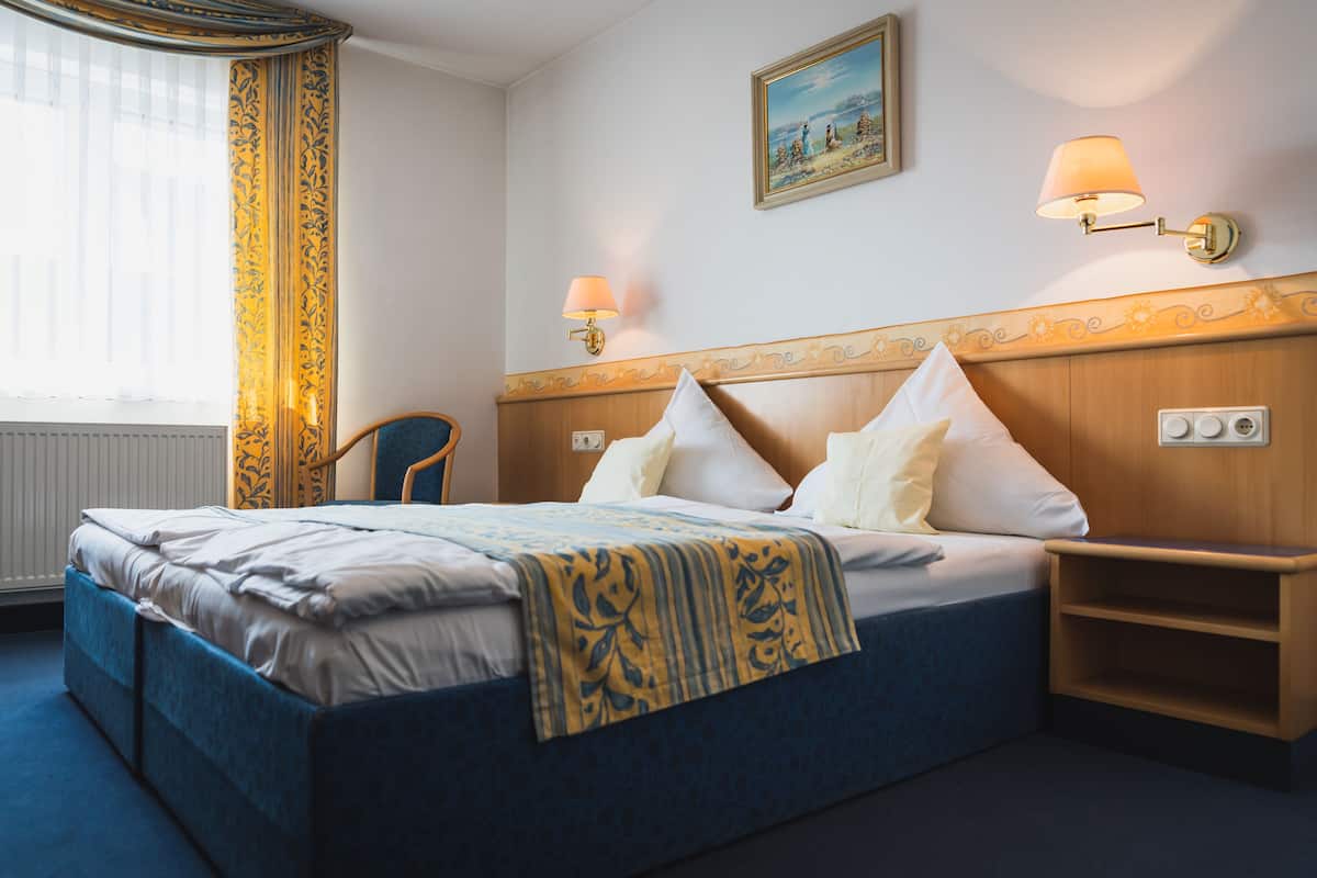 Quarto de hotel com uma cama grande, duas luminárias de cabeceira, um painel de cabeceira em madeira, um quadro na parede e uma janela com cortinas.