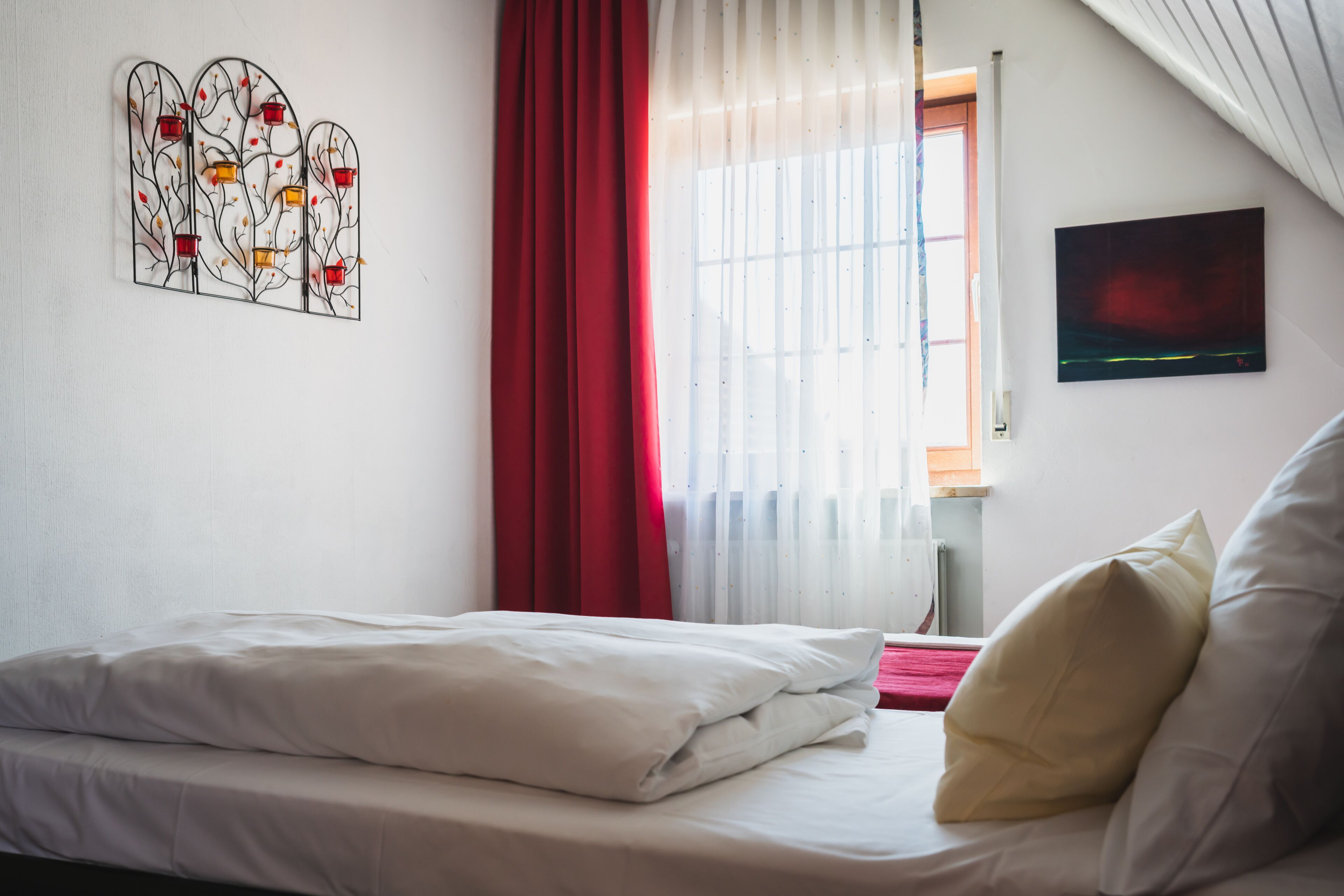 Un dormitorio con cama, ropa de cama blanca, una cortina roja, una ventana con cortinas traslúcidas y una obra de arte colgada en la pared.
