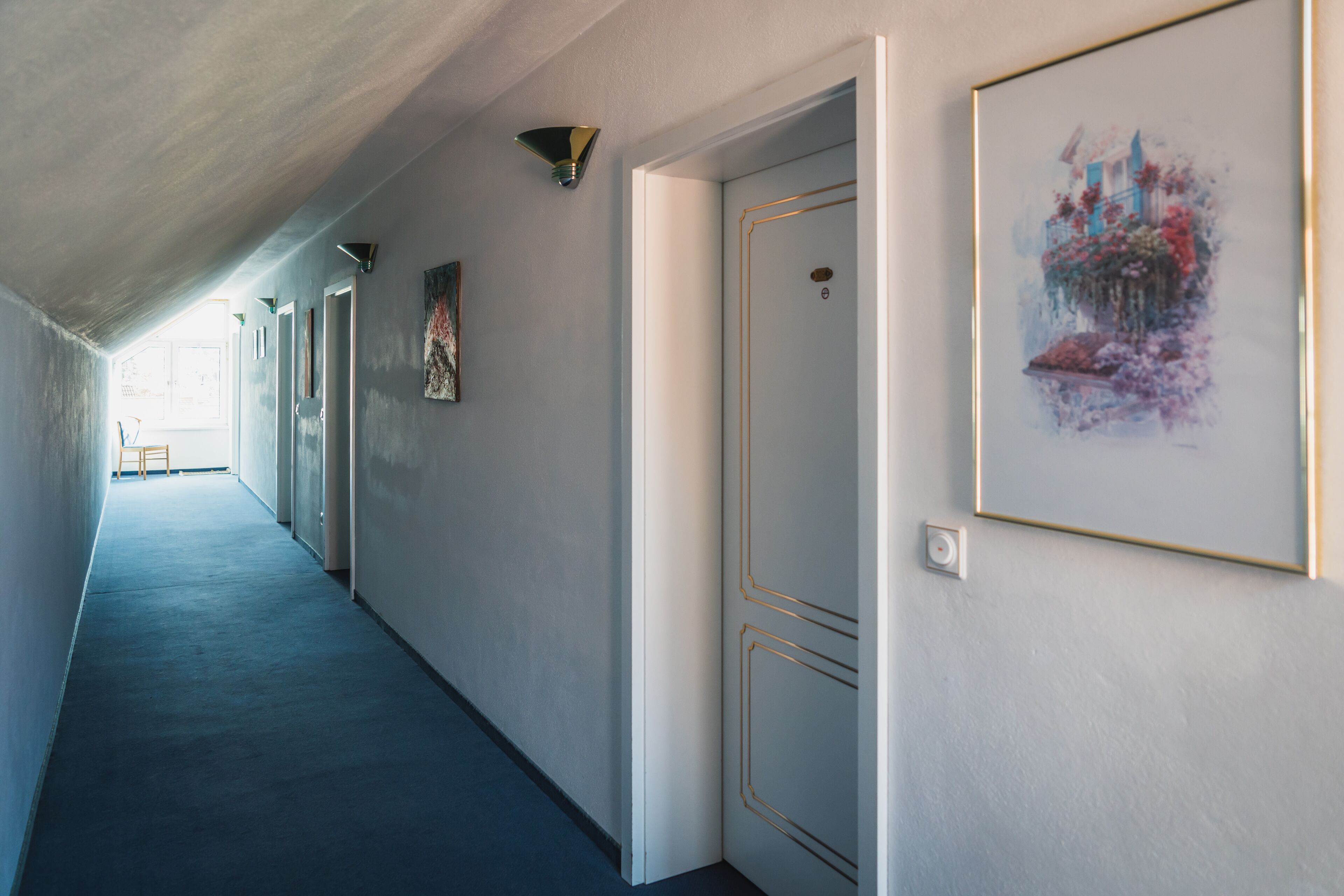 A hallway with a blue carpet, white walls, and a door with a gold frame. There are framed artworks on the walls and a light fixture above the door.