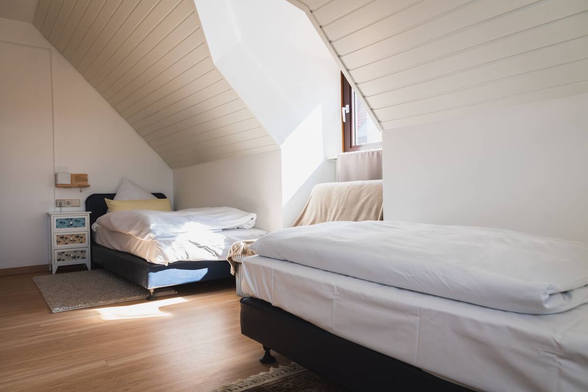 Quarto com teto inclinado, duas camas, uma mesa de cabeceira e uma janela.