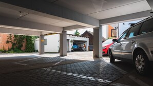 Et overdækket parkeringsområde med belagt gulv, en hvid bil parkeret til højre og et blåt køretøj i baggrunden.