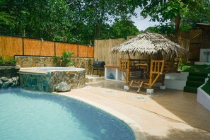 A swimming pool with a hot tub, a wooden deck, and a thatched-roof seating area.